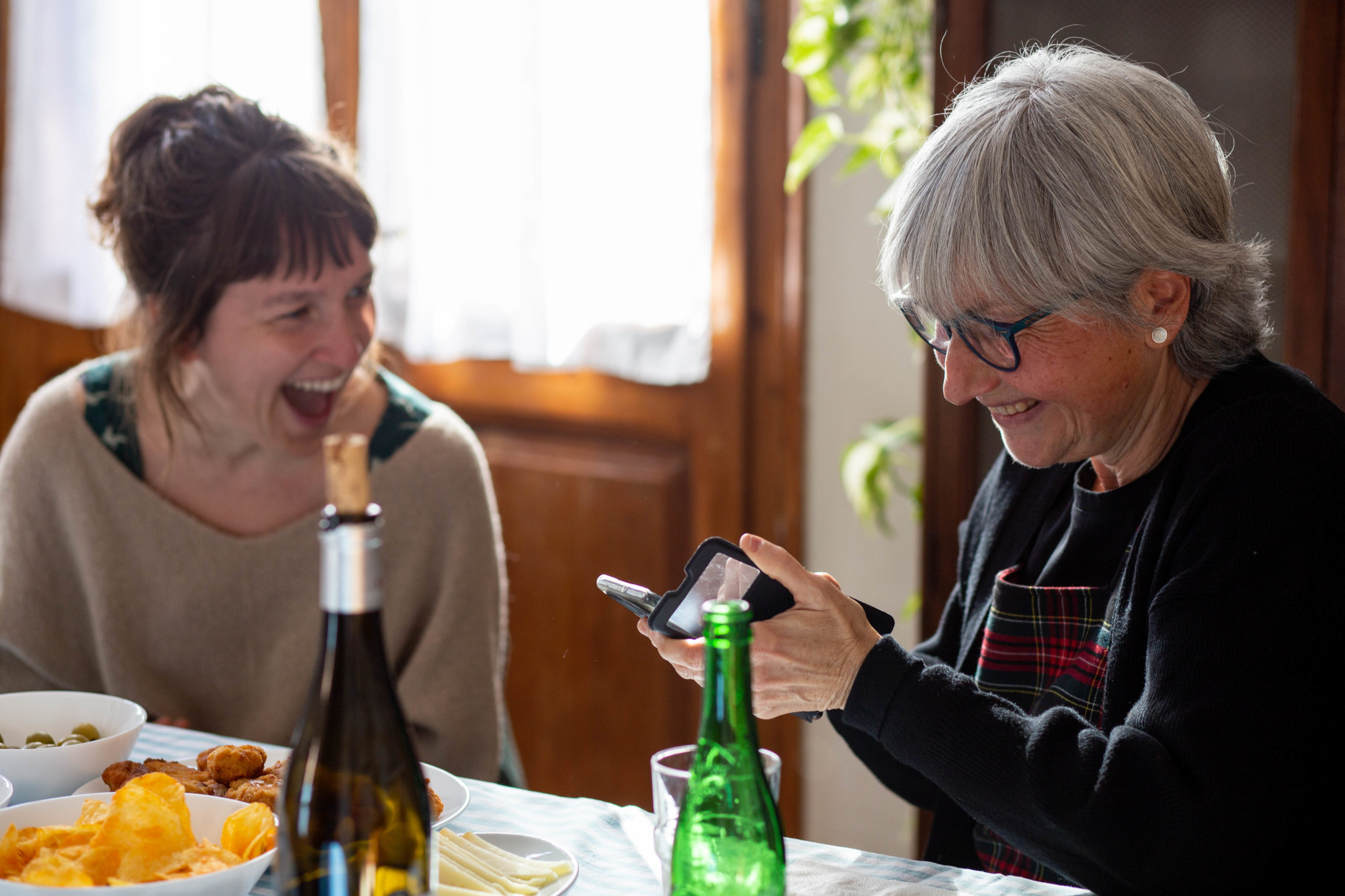 Dues amigues rient en un dinar mentre miren el mòbil en un menjador de casa