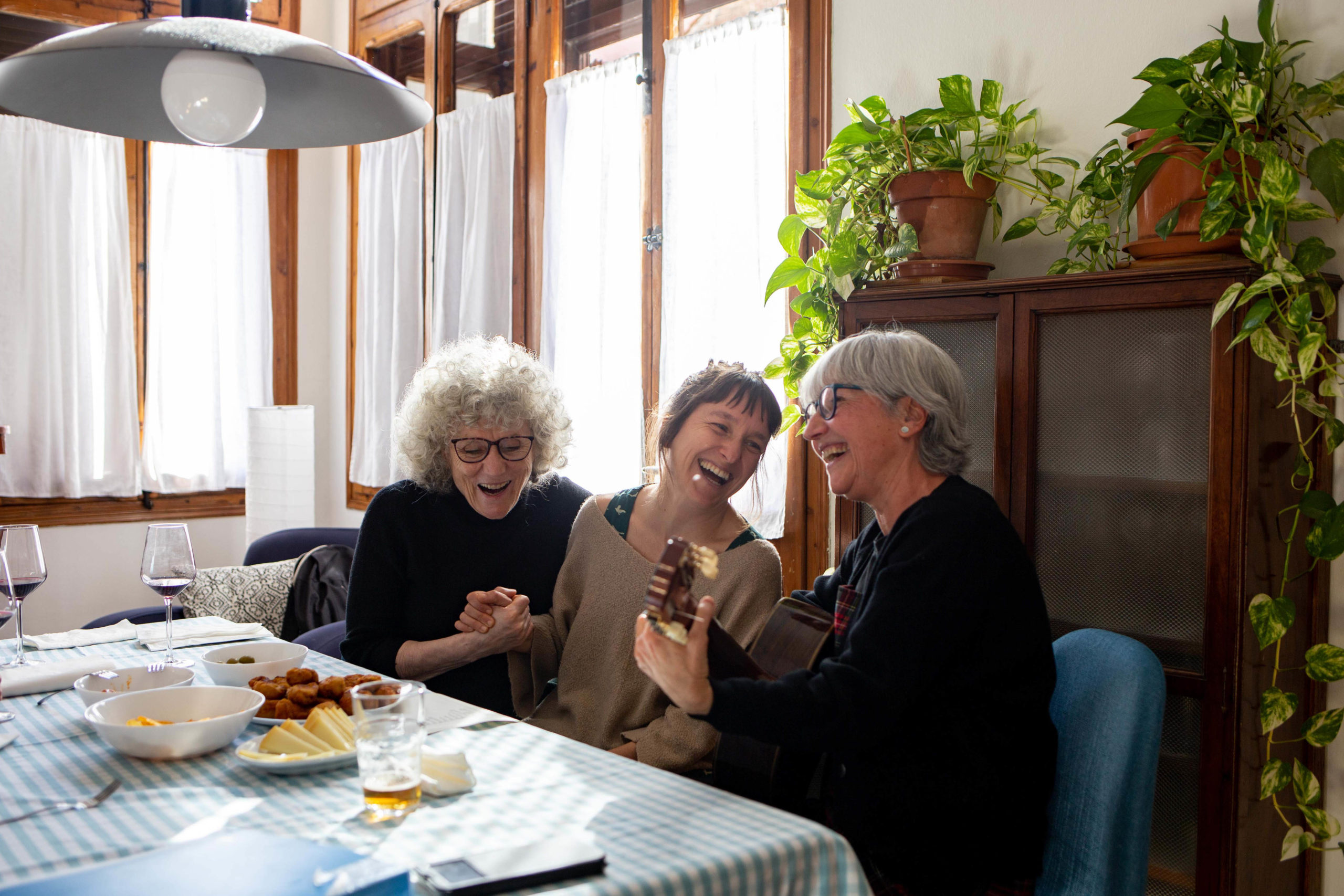 Tres amigues tocant la guitarra i rient al menjador de casa